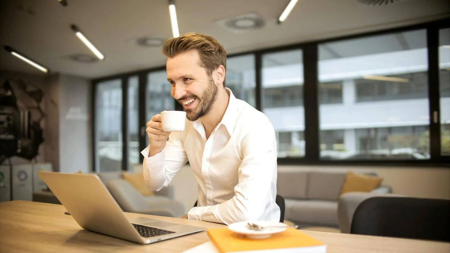 Muž pracuje na počítači, usmívá se a pije kávu