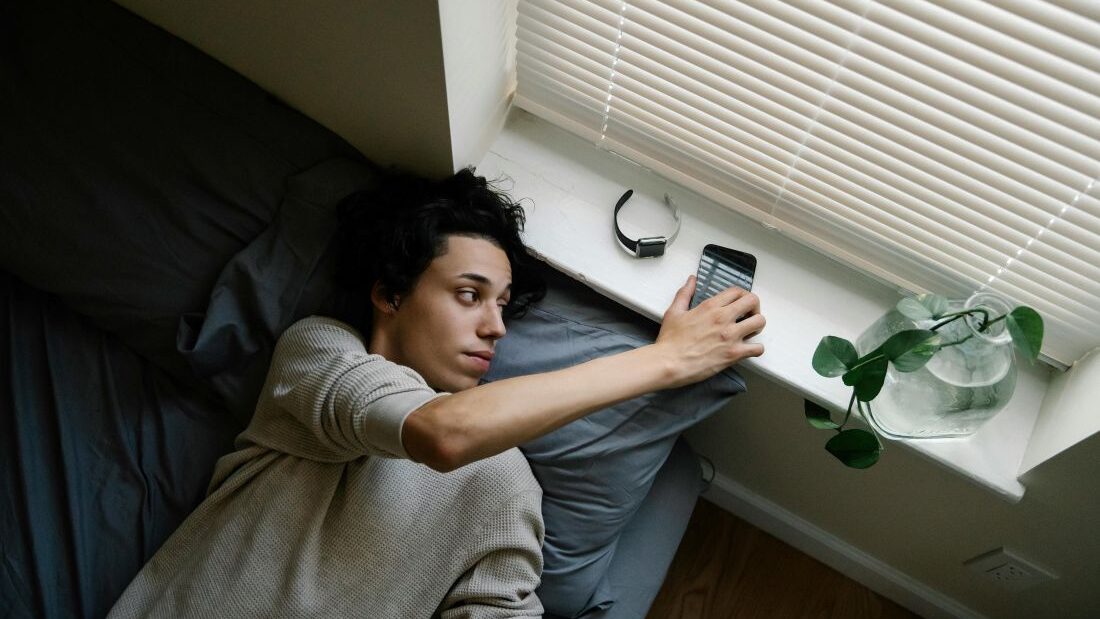 Muž ráno leží v posteli a natahuje se na parapet pro telefon.