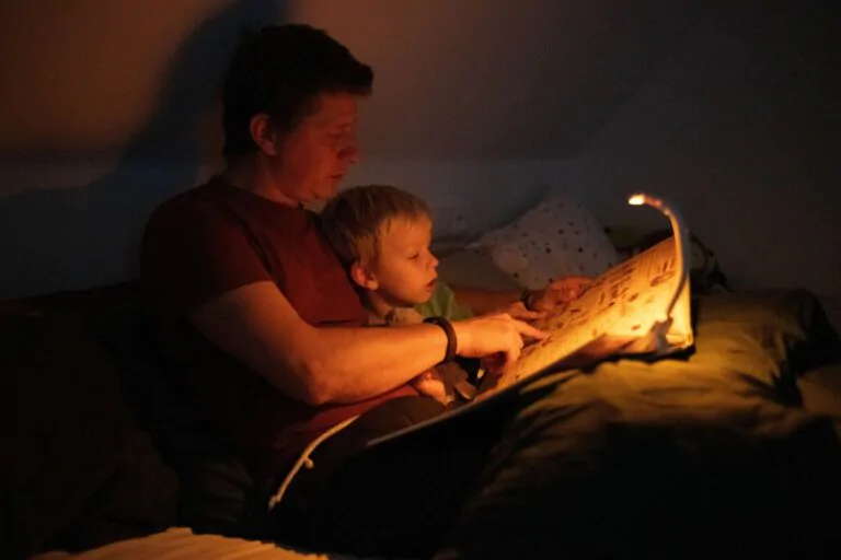 Tatínek s dítětem večer leží v psoteli s čtou si pohádku a svítí jim večerní LED lampička s klipem Oranžovka.