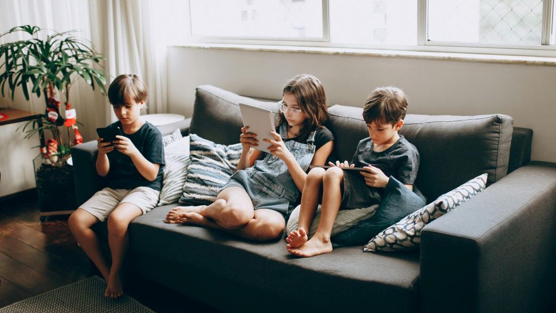 Tři děti sedí na gauči a sledují mobily a tablety.