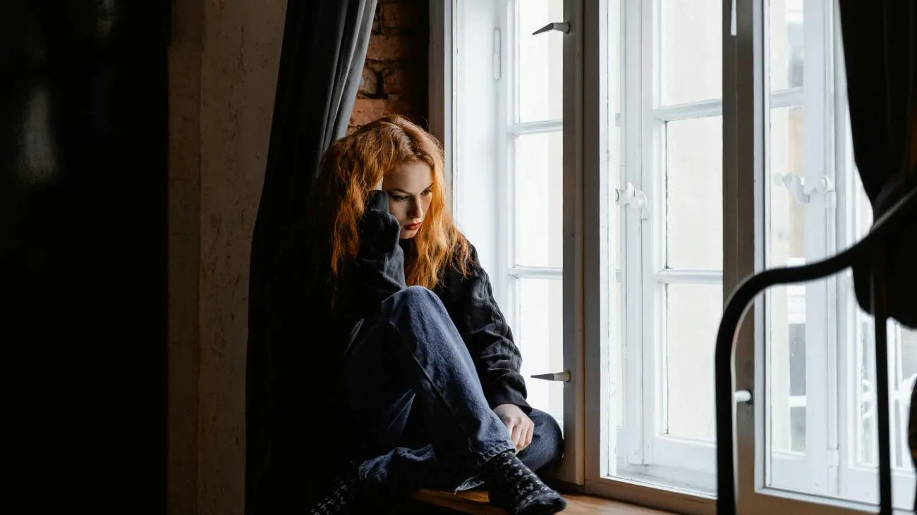 Žena sedí na parapetě okna, dívá se ven a má špatnou náladu.