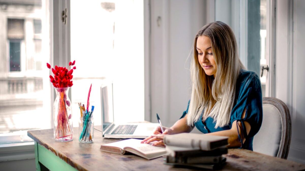 Žena sedí u stolu blízko okna a pracuje.