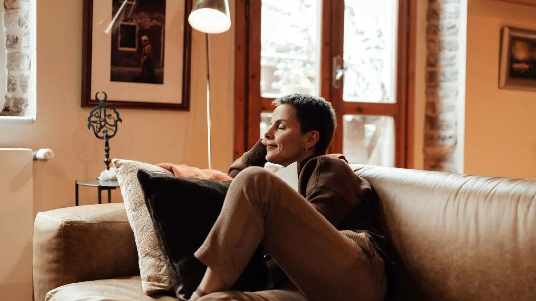 Žena sedí na pohovce pod oranžovým světlem stojací lampy a relaxuje se zavřenýma očima.